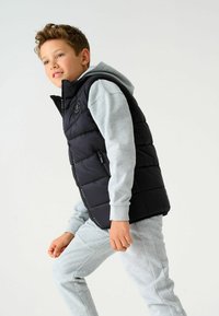 Melna pūkainā veste ar augstu apkakli, ar rāvējslēdzēja kabatām un logo detaļām, valkāta virs pelēka kapucīna un saskaņotiem sporta biksēm. Gluda tekstūra.