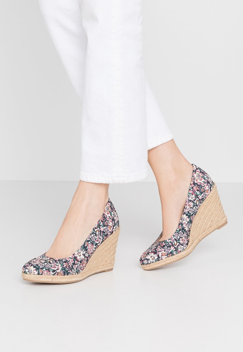 Femme portant des chaussures compensées à motif floral avec talons tissés beige et pantalon blanc court, debout sur un sol clair uni.