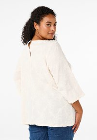 Blusa beige chiaro con ricamo floreale testurizzato, scollo rotondo, maniche a 3/4 e una piccola apertura sul retro, abbinata a jeans di denim scuro.