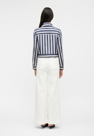 Γυναίκα που στέκεται γυρισμένη πλάτη, φοράει ένα μπλε και άσπρο σακάκι με κατακόρυφες ρίγες, άσπρο παντελόνι με φαρδιά μπατζάκια και μαύρα παπούτσια με τακούνι.