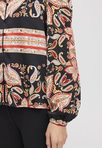 Blouse noire ornée de motifs paisley et floraux colorés, dotée d'une fermeture par boutons et de poignets froncés. Texture soyeuse, éléments de design audacieux.