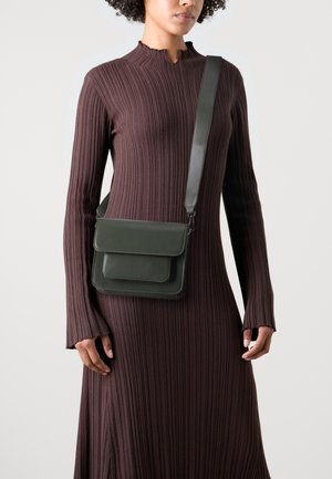 Sac bandoulière en cuir vert avec deux rabats avant, texture lisse et sangle réglable. Porté avec une robe brune à manches longues côtelée.