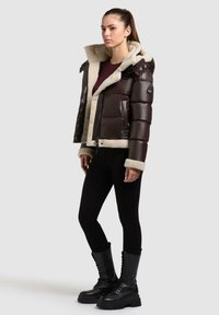 Braune gepolsterte Jacke mit einem Schaffellkragen, Reißverschluss und schwarzen Akzenten, kombiniert mit schwarzen Leggings und knielangen geschnürten Stiefeln.