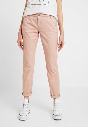 Personne portant un pantalon rose clair avec revers, des baskets montantes blanches et un t-shirt blanc avec un graphisme noir sur un fond blanc uni.