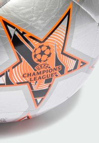 Pallone da calcio con una superficie argentata decorata con motivi di stelle arancioni e nere, logo della UEFA Champions League e pannelli testurizzati per una presa migliore.