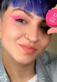 Rosa kosmetikkbeholder merket "371" holdt av en person med lilla hår, iført en dongerijakke. Merkbar eyeliner og subtil leppefarge.