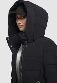 Sort pufferjakke med en stor, struktureret hætte. Funktioner inkluderer trykknapper, lynlås foran og en glat, quilted tekstur.