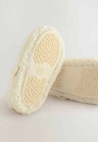 Vita, fluffiga tofflor med en texturerad, beige gummisula som har ett blommönster, märkta "NEXT," och med mjukt, lurvigt material på ovansidan.