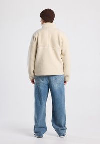 Giacca in pile crema con colletto alto, abbinata a jeans in denim blu larghi. Presenta un design semplice e pulito e una texture leggera.