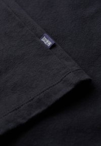 Camiseta de algodón negra con una manga doblada y una pequeña etiqueta azul. La tela tiene una textura suave y costuras visibles.