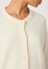 Light cream cardigan with a ribbed neckline, button closure, soft texture, and a relaxed fit. Features subtle fabric variations for added detail.