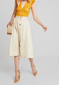 Spetsbodysuit i klar orange med volangärmar, kombinerad med en krämfärgad midikjol med knappar och nakna ankelremmar. Vävda handväska ingår.