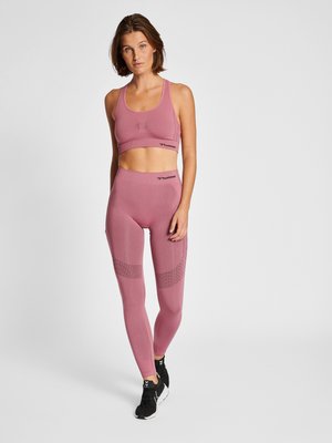Kvinde stående iført mauve sømløs sports-bh og leggings med sorte sportssko på hvid ens baggrund.