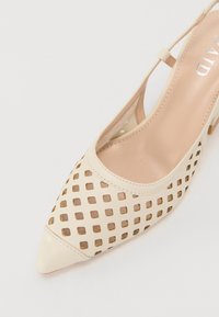 Chaussure slingback beige à bout pointu avec motif découpé carré et talon bas sur fond blanc.