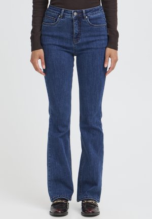 Femme portant un jean bootcut bleu foncé, un haut noir à manches longues et des mocassins noirs avec un détail chaîne dorée, debout devant un fond uni.