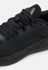 Chaussures de sport noires avec un dessus texturé, fermeture à lacets et une marque tonale bien visible. La semelle extérieure présente un profil de bande de roulement pour une meilleure adhérence.