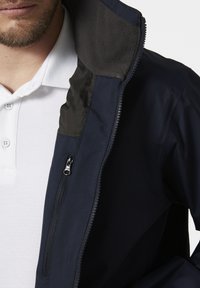 Marineblauwe softshell jack met een grijze fleece-voering in de kraag, rits-sluiting en een zijzak met rits. Gekozen over een wit overhemd met kraag.