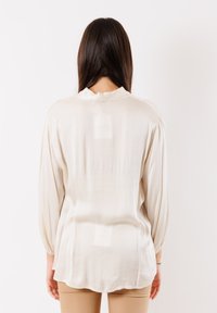 Blusa color crema con una texture liscia e setosa, scollatura rotonda e maniche lunghe arricciate, caratterizzata da una leggera lucentezza e un orlo dritto.