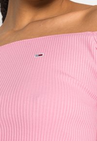 Růžový žebrovaný top s odkrytými rameny, který obsahuje malou výšivku s logem. Látka je elastická a má hladkou strukturu.