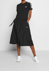 Person som bär en svart Adidas kortärmad t-shirt och culottes med vita ränder, händerna i fickorna, och vita sneakers mot en enkel bakgrund.