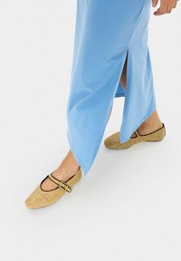 Ballerine beige intrecciate con punta arrotondata, rifiniture nere e cinturino regolabile, abbinate a un lungo vestito azzurro chiaro con uno spacco laterale.