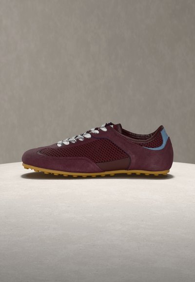 Κρυφή μπορντό αθλητική παπούτσια με πλαινά πάνελ από δίχτυ, λευκά κορδόνια, γαλάζια λεπτομέρεια στη φτέρνα και κίτρινη ελαστική σόλα σε μπεζ επιφάνεια.