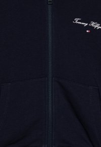 Mørkeblå zip-up hettegenser med frontlommer, glidelås, og en liten hvit "Tommy Hilfiger" logo med rødt, hvitt og blått flaggdetalj.