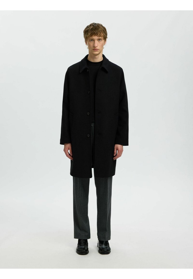Jeune homme debout portant un manteau noir boutonné, un pull foncé, un pantalon gris et des chaussures élégantes noires sur un fond clair uni.
