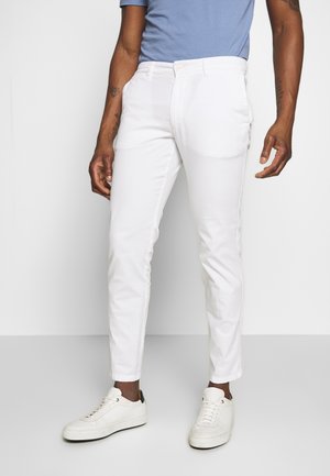 Mężczyzna w białych spodniach o dopasowanym kroju, jasnoniebieskiej koszuli i białych trampkach, stojący na tle jednolitego szarego tła.