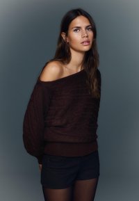 Dark brown cable knit sweater with an off-shoulder design, featuring wide sleeves and a ribbed hem. Paired with dark shorts.