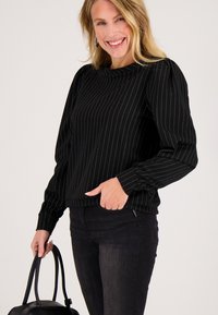 Zwarte trui met witte verticale strepen, lange mouwen met gerimpelde manchetten, ronde halslijn en een losse pasvorm. Gecombineerd met zwarte jeans.