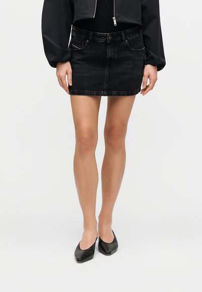 Mini-jupe en denim noir à coupe droite, avec poches avant et fermeture par boutons, associée à des chaussures noires à bouts pointus.