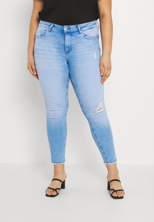 Femme portant un jean skinny déchiré bleu clair avec un ourlet brut et des sandales à talons blocs noires à brides sur fond blanc.