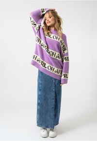 Pull en tricot violet surdimensionné avec le texte "APRIL" en rayures noires, associé à une longue jupe en denim avec des boutons et des chaussures à plateforme blanches.