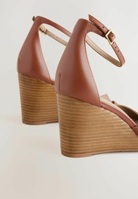 Zuecos de cuña de cuero marrón con una suela de madera texturizada y correa ajustable en el tobillo, que cuentan con un acabado suave y un diseño de puntera redondeada.