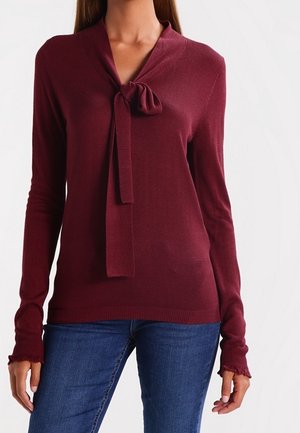 Pull en bourgogne à manches longues avec un nœud noué au niveau du col. Poignets et ourlet côtelés, tissu en tricot texturé, associé à un jean bleu.