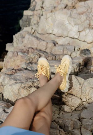 Baskets en toile jaunes avec des semelles en caoutchouc blanches et des pointes arrondies, équipées de lacets et d'œillets métalliques, posées sur un terrain rocheux.