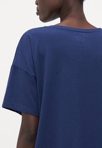 Námorníckomodrá tričko s krátkymi rukávmi, širokým výstrihom a jemne embosovaným logom v oblasti krku vzadu. Mäkký bavlnený materiál, voľný strih.