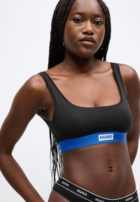 Musta rintaliivit sileästä kankaasta, joissa on suorakaiteen muotoinen pääntie ja leveä sininen joustava nauha, jossa on "HUGO"-logo. Mallilla on lettipäinen kampaus.