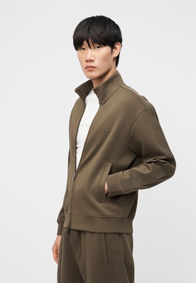 Νέος άνδρας που φοράει ελιάς πράσινο μπουφάν με φερμουάρ και ταιριαστό παντελόνι, στέκεται με το ένα χέρι στην τσέπη σε απλό φόντο.