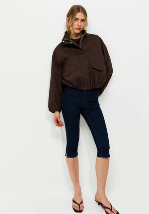 Donna con giacca marrone scuro con cerniera, jeans skinny blu scuro corti e sandali neri con tacco, in piedi davanti a uno sfondo bianco.