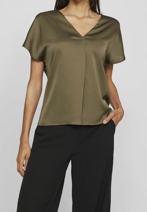 Kvinde iført olivengrøn kortærmet satinbluse med V-udskæring og sorte højtaljede bukser, hånd i lomme, mod ensfarvet grå baggrund.