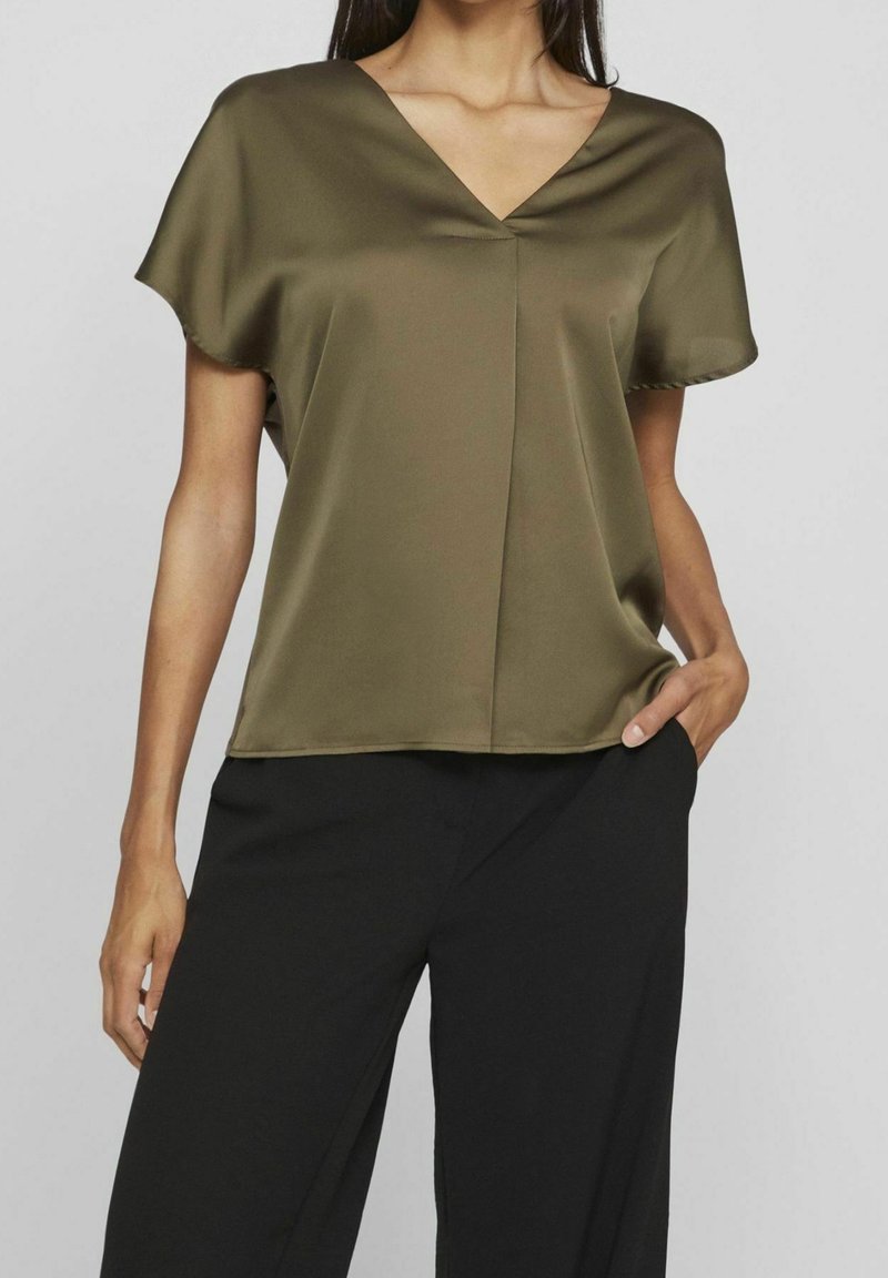 Femme portant un chemisier en satin vert olive à manches courtes avec col en V et un pantalon noir taille haute, main dans la poche, devant un fond gris uni.