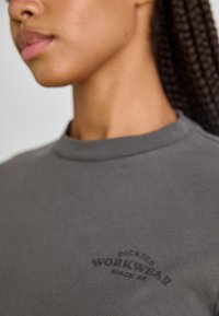 Pilkas megztinis, pagamintas iš minkštos medžiagos, su iškilnia apykakle ir juodu spausdintu logotipu "DICKIES WORKWEAR SINCE 1922."