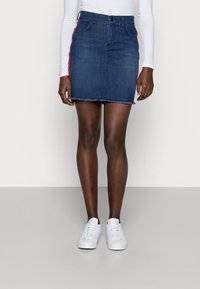 Džínová mini sukně v tmavě modré barvě s červenými postranními pruhy, s potrhaným lemem a tradičním designem s pěti kapsami, v kombinaci s bílými teniskami.