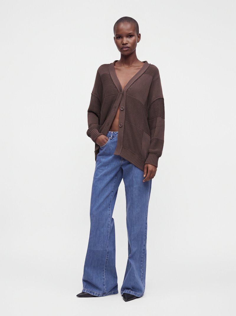 Modèle portant un cardigan en maille marron oversize avec deux boutons, un jean large bleu et des chaussures noires à bout pointu, debout sur fond blanc.