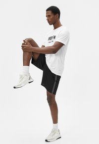Hvid sports t-shirt med sort grafisk tekst, parret med sorte shorts med sideløse striber, og hvide sports sneakers med grå accenter.