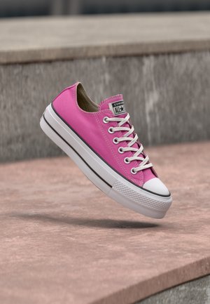 Jasnoróżowy niski sneaker Converse z białymi sznurowadłami i grubą białą podeszwą unoszący się nad kamienną powierzchnią.