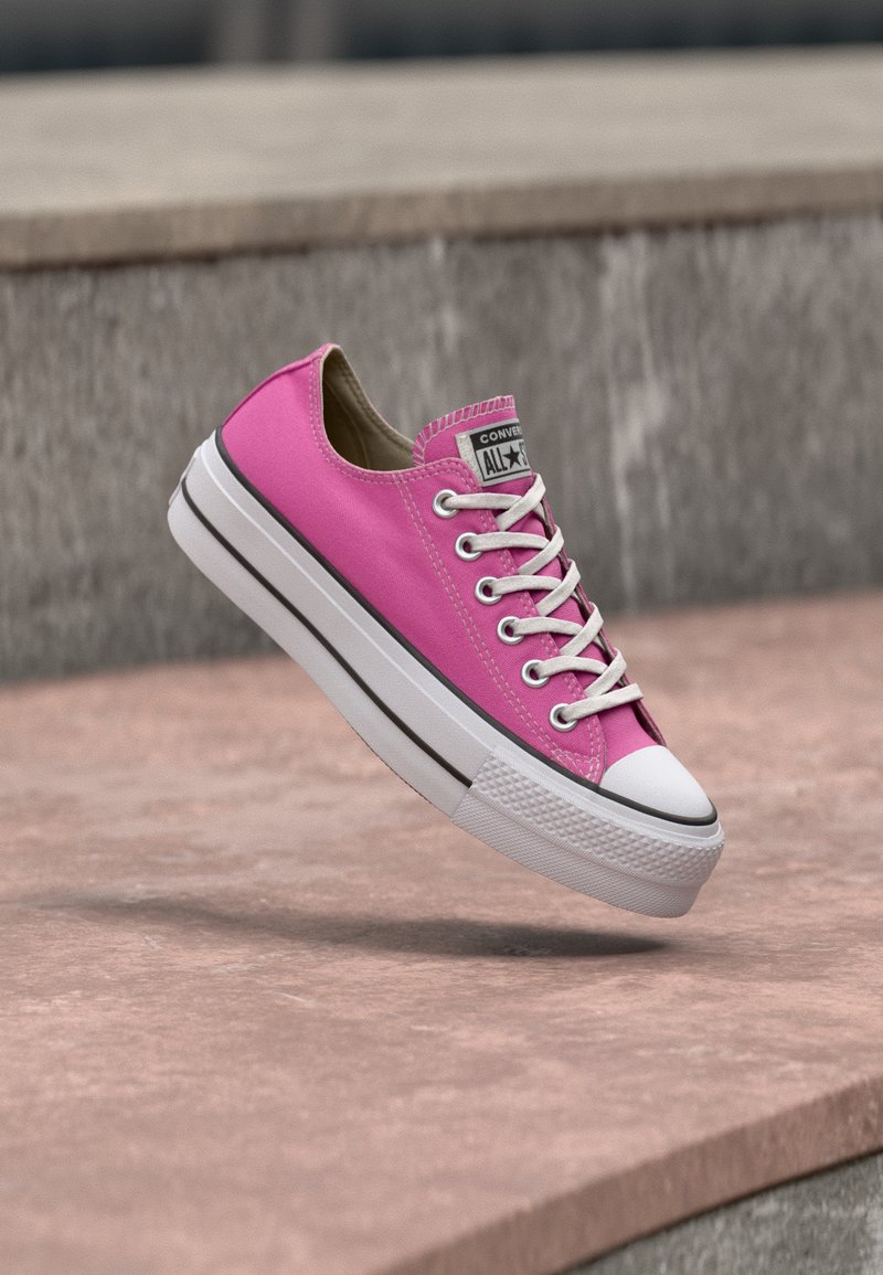 Φωτεινό ροζ χαμηλό παπούτσι Converse με λευκά κορδόνια και χοντρή λευκή σόλα, αιωρούμενο πάνω από μια πέτρινη επιφάνεια.