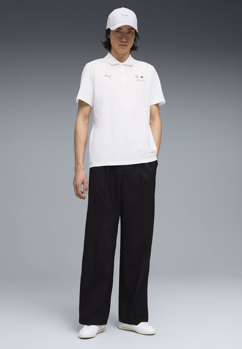 Jeune homme debout, portant un polo et une casquette Puma Motorsport blancs, un pantalon large noir et des baskets blanches, sur fond gris.